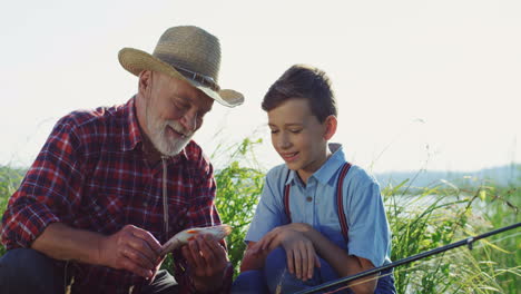 viejo abuelo y nieto sentados en la orilla del río y mirando los peces que atraparon