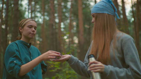 two friends share peaceful moment in forest as one pours warm drink from thermos, her friend smiles and waits to receive it, surrounded by tall trees, they enjoy a serene outdoor break together