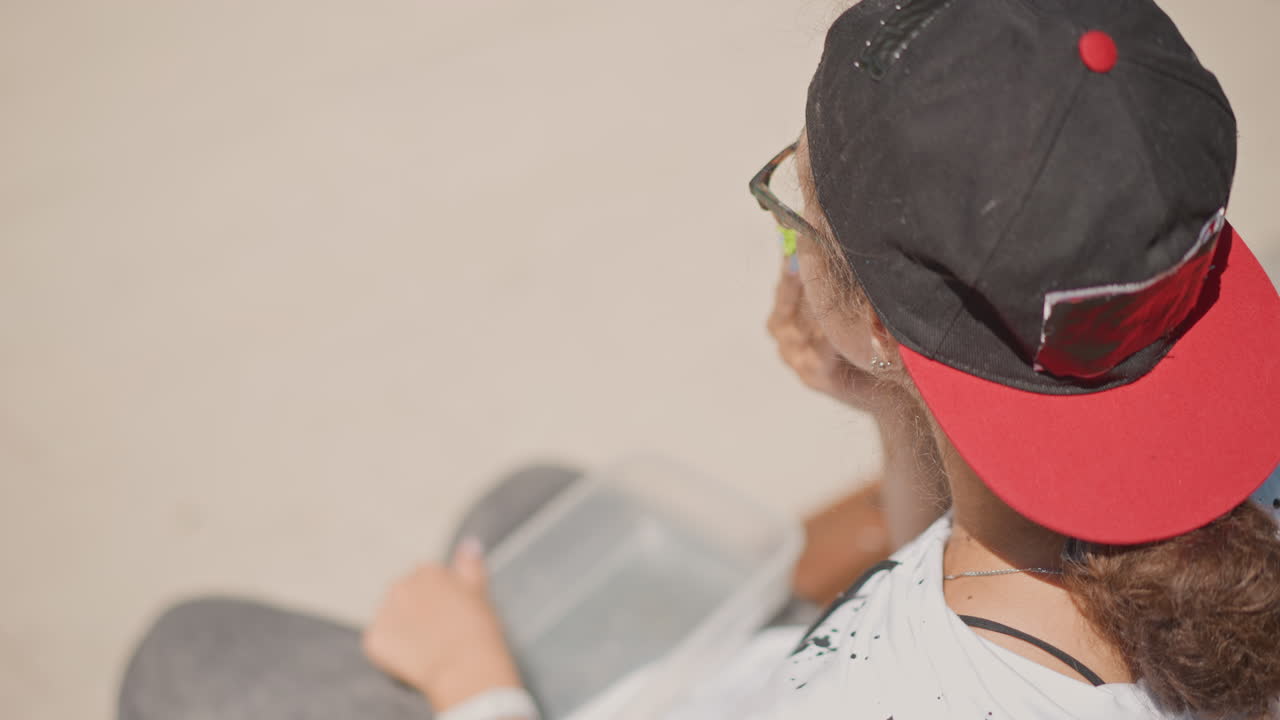 Mujer joven con monopatín sentada al aire libre con gorra roja al revés y gafas, pelo rizado recogido en un moño bajo, sujetando el teléfono en el regazo, tabla de monopatín al lado, enfoque superficial cenital, tonos cálidos beige
