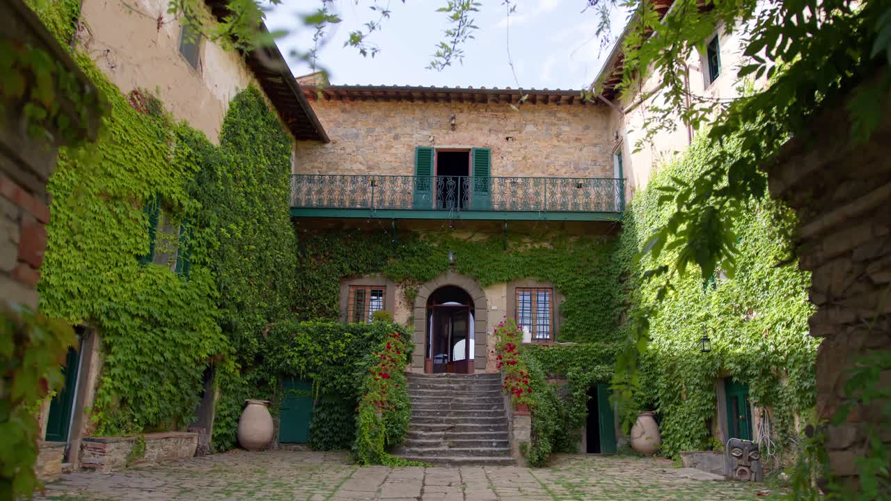 Ampia ripresa al rallentatore di una passeggiata attraverso un arco di porta verso una villa toscana, con piante mosse dal vento, luce solare e calde atmosfere estive