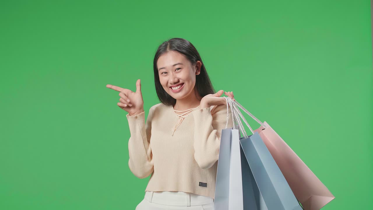 An Asian Shopping Woman Holding Shopping Bags Up Before Pointing And Smiling To Camera While Standing In Front Of Green Screen