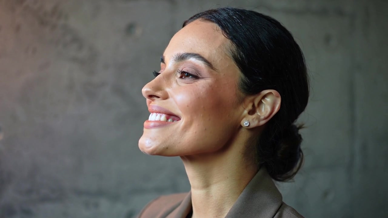 Donna sorridente in blazer marrone che alza il mento dopo l'indicazione della telecamera sul set, con orecchini a perno e spazio per il testo.
