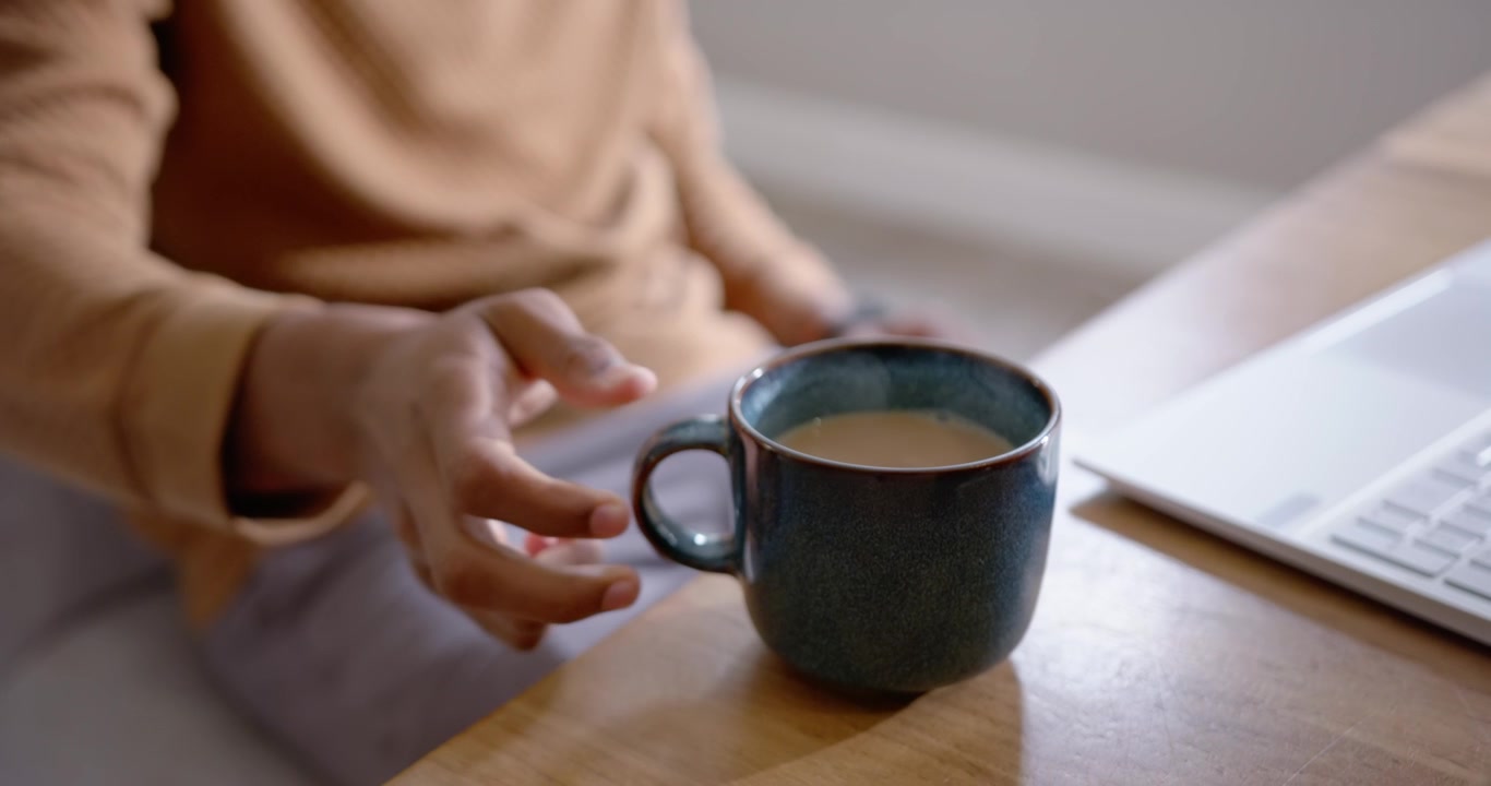 Uomo afroamericano che sposta in avanti una tazza di ceramica blu su un tavolo di legno, digitando al laptop.