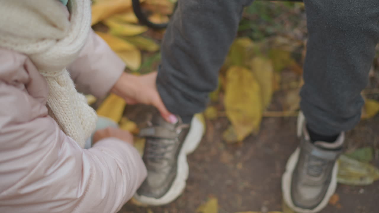 close up top down view of mother crouching over child foot adjusting shoe strap on sneaker over fallen autumn leaves near bench iron work by tree trunk conveying nurturing care