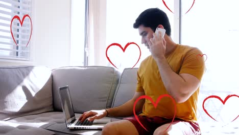 Multiple-red-heart-icons-falling-over-caucasian-man-using-laptop-and-talking-on-smartphone-at-home