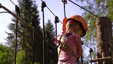 niña europea de 6 años pasando un puente de madera en la copa de un árbol dentro del parque de escalada voss - actividad de vacaciones de verano con equipo de seguridad y hermosa luz del sol que viene desde atrás - cámara lenta noruega