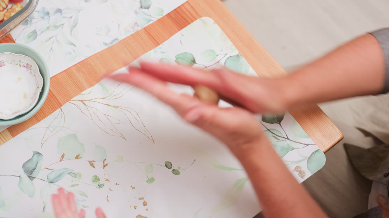 Close up of adult hands rolling dough on mat, dropping piece then rolling another, second hands partially visible during classroom activity, showing technique, guidance and cooking prep