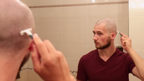 young bald man shaving his own head while looking in a mirror, view of back but focus on reflection