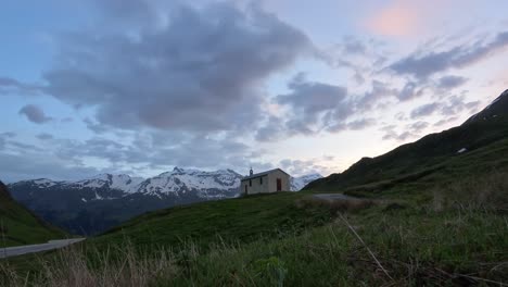 Zeitraffer-Ansicht-Eines-Einsamen-Hauses-Auf-Einem-Grünen-Hügel-Unter-Einem-Weiten-Himmel-Mit-Schneebedeckten-Bergen-Im-Hintergrund,-Was-In-Der-Abenddämmerung-Eine-Ruhige-Und-Malerische-Szenerie-Schafft