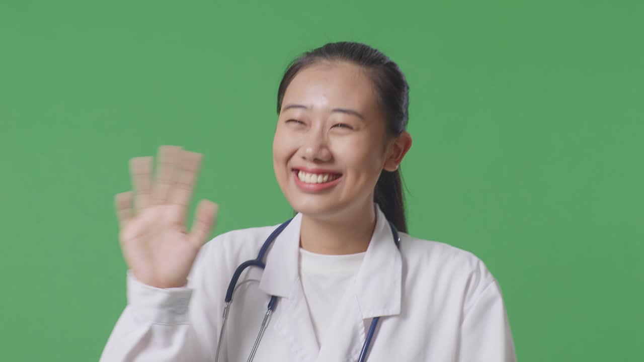 스테토스코프를 들고 있는 아시아 여성 의사의 근접 사진. 손을 흔들고 미소 짓고 병원에서 녹색 화면 배경에서 고 있다.