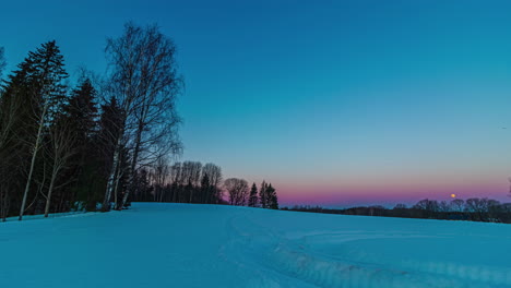 Timelapse-De-La-Hora-Azul-Sobre-El-Campo-Cubierto-De-Nieve-Con-árboles-Y-Luna-Puesta-En-El-Cielo-En-La-Distancia