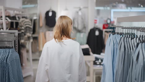 back view of woman walking through clothing store surrounded by racks of blue and patterned shirts, bright lighting and organized setup create a modern shopping environment