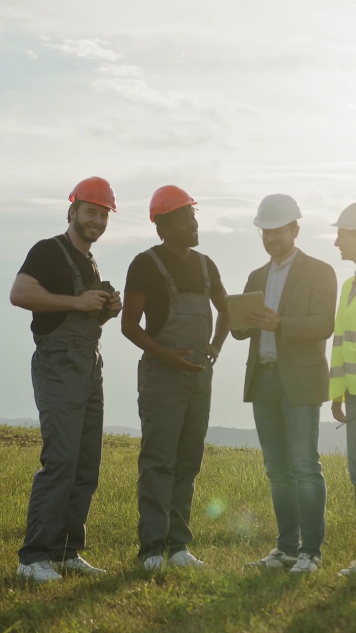 Diverse construction workers in hard hats and safety vests gather outdoors discussing plans on a