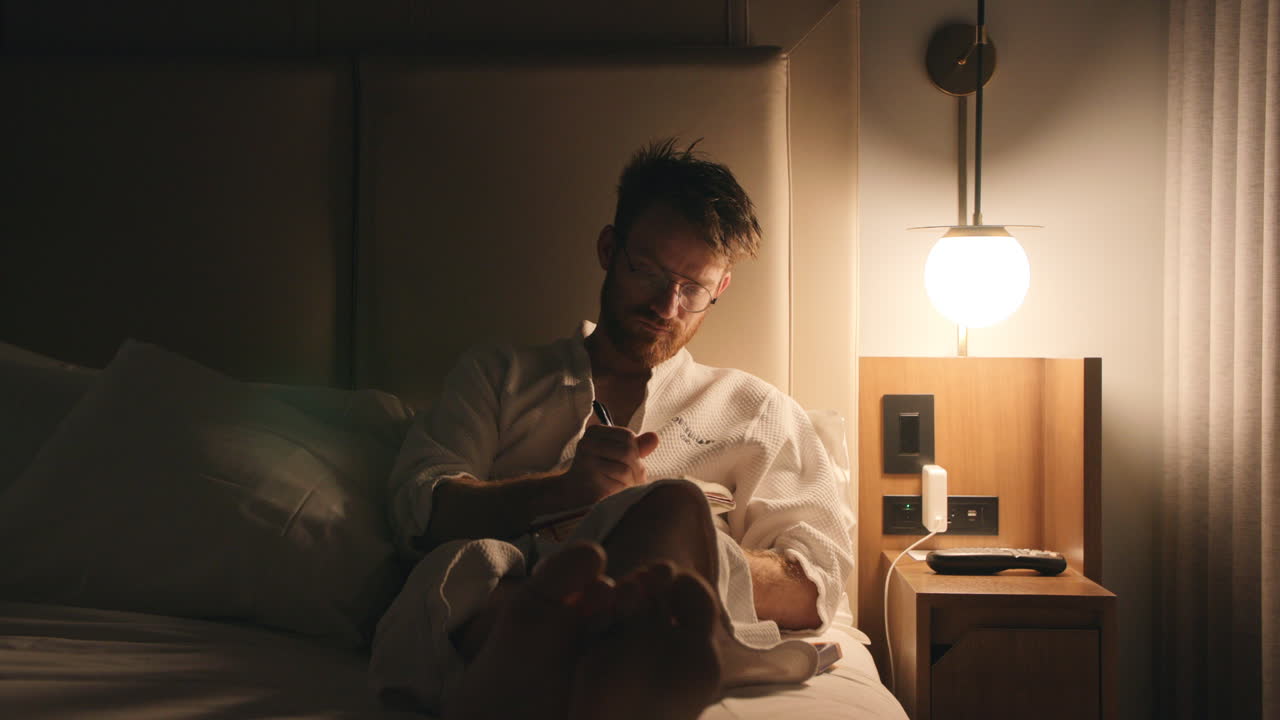 Red haired man in bed with a robe on writing in a journal.