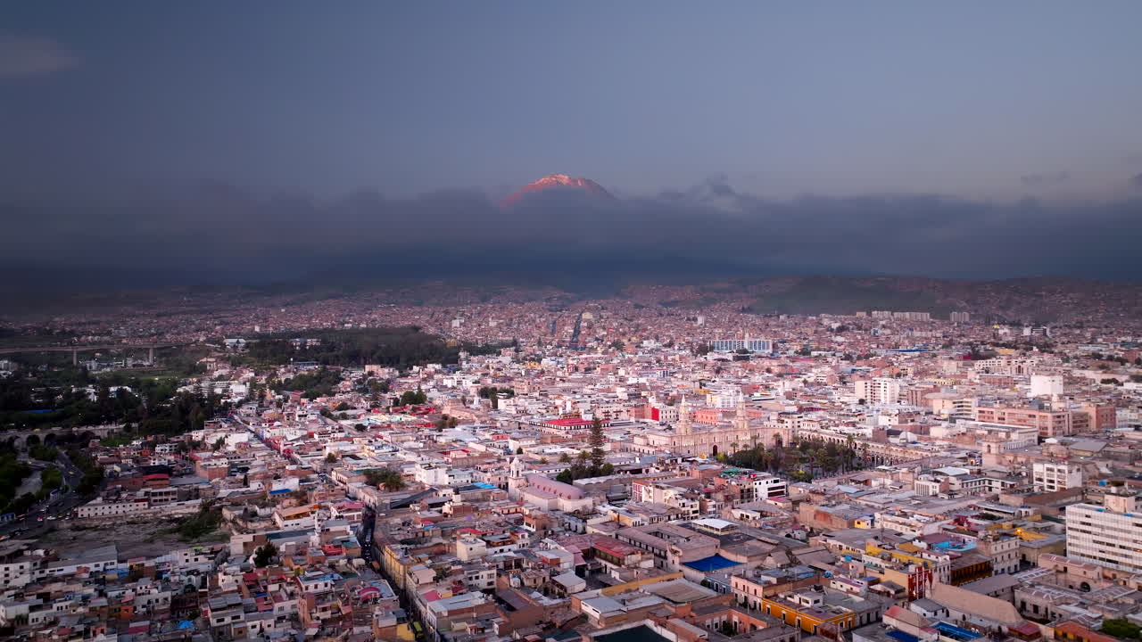 Vista aérea cinematográfica de Arequipa, Perú, al atardecer. Denso paisaje urbano bajo una capa de nubes sombrías con un pico volcánico en el horizonte en América del Sur.