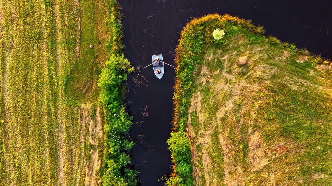 Top view of a rowing boat sailing on river surrounded by green grassy banks. Drone shot