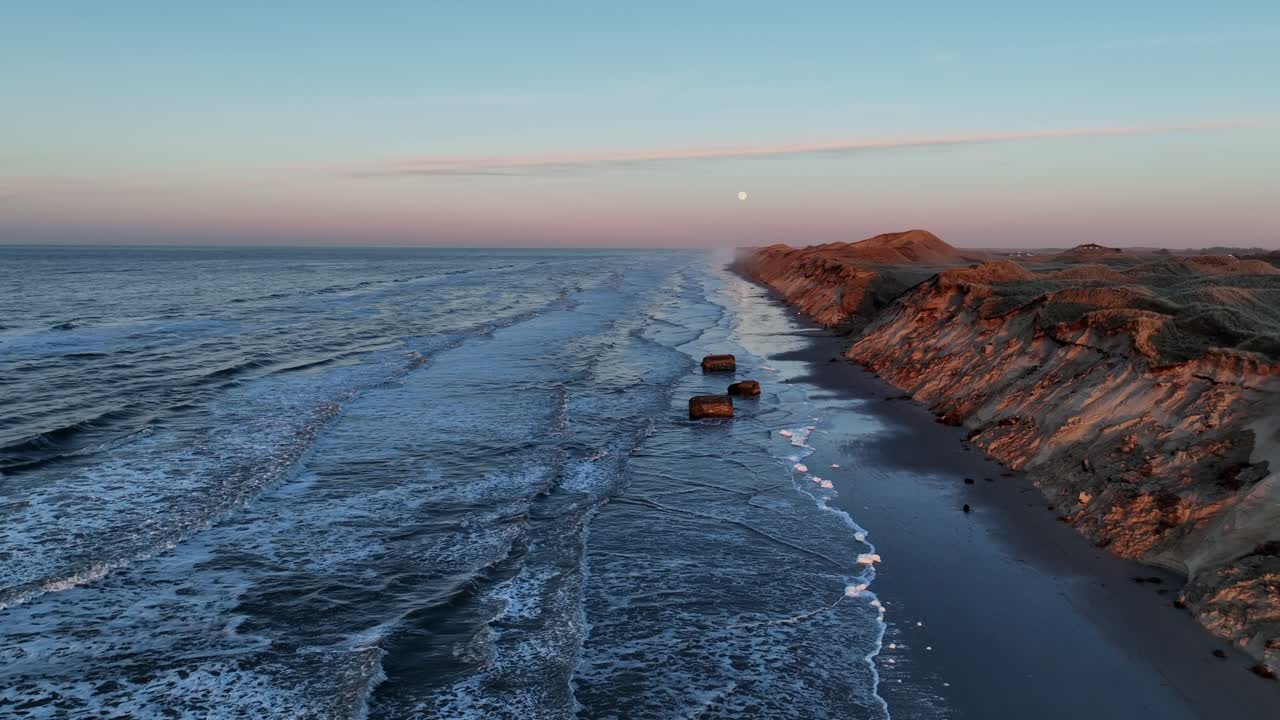vista aérea las olas del mar están lavando en la playa, altas dunas se han formado, los restos de bunkers de guerra están en la costa, y el sol se está poniendo en una noche de invierno