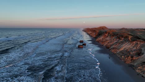 vista aérea las olas del mar están lavando en la playa, altas dunas se han formado, los restos de bunkers de guerra están en la costa, y el sol se está poniendo en una noche de invierno