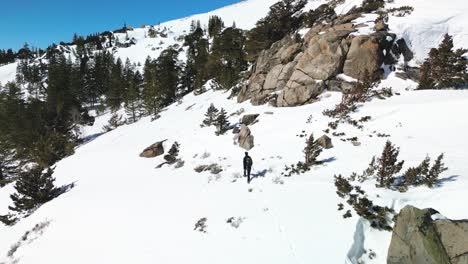 Aerial-view-of-hiker-walking-in-deep-snow-along-rock-cliffs,-Carson-Pass,-California