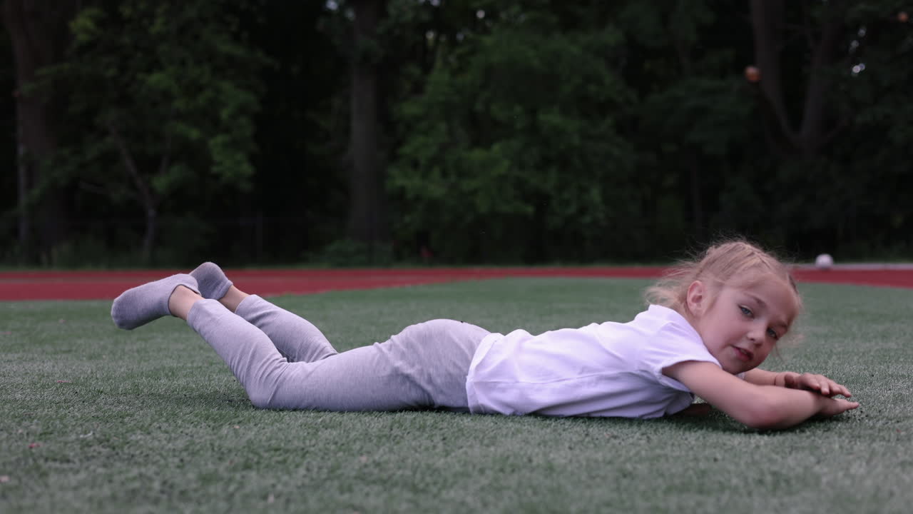 A little girl lies on the green grass and looks at the camera. A stylish girl in a white T-shirt. A girl with braided hair