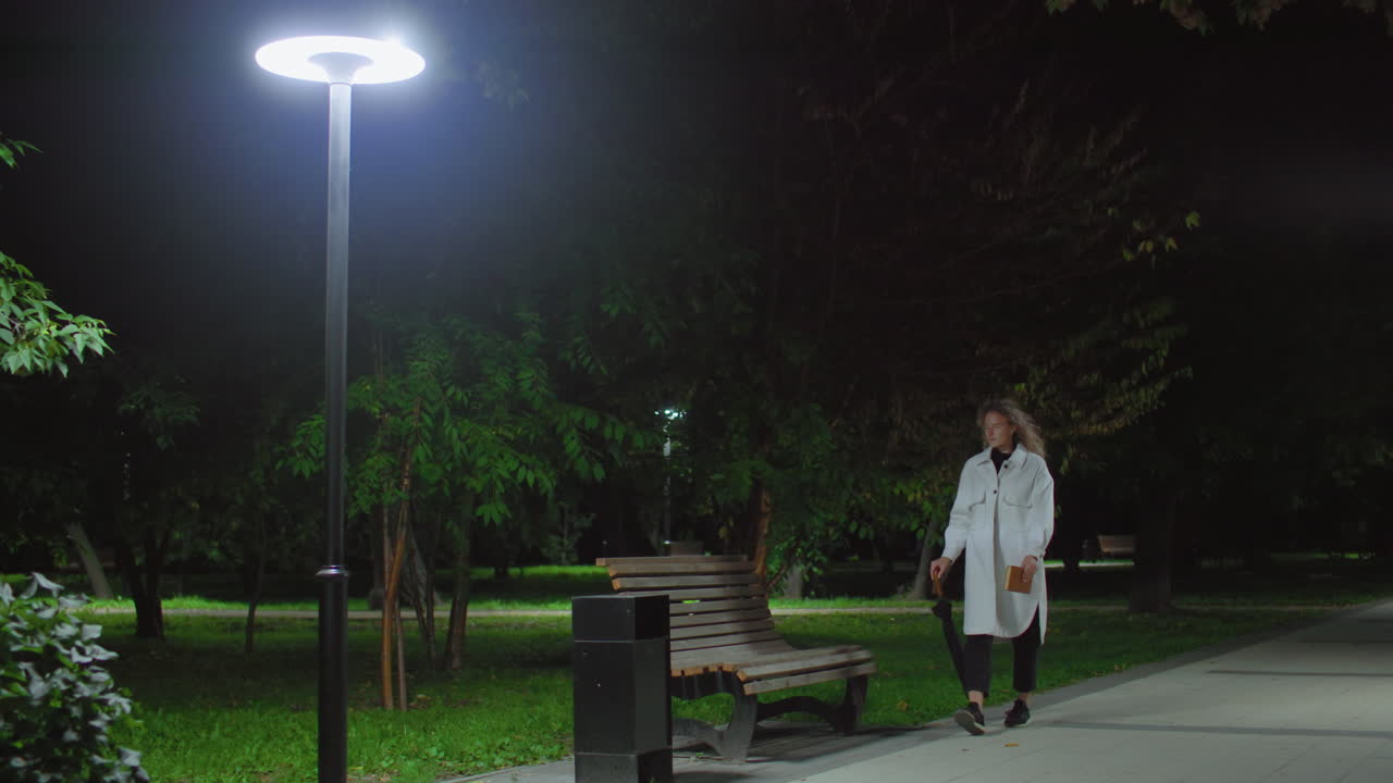 Novelist walks toward wooden bench holding umbrella and book under glowing streetlight, surrounded by trees swaying gently in wind, prepares to sit, cross legs, adjust hair