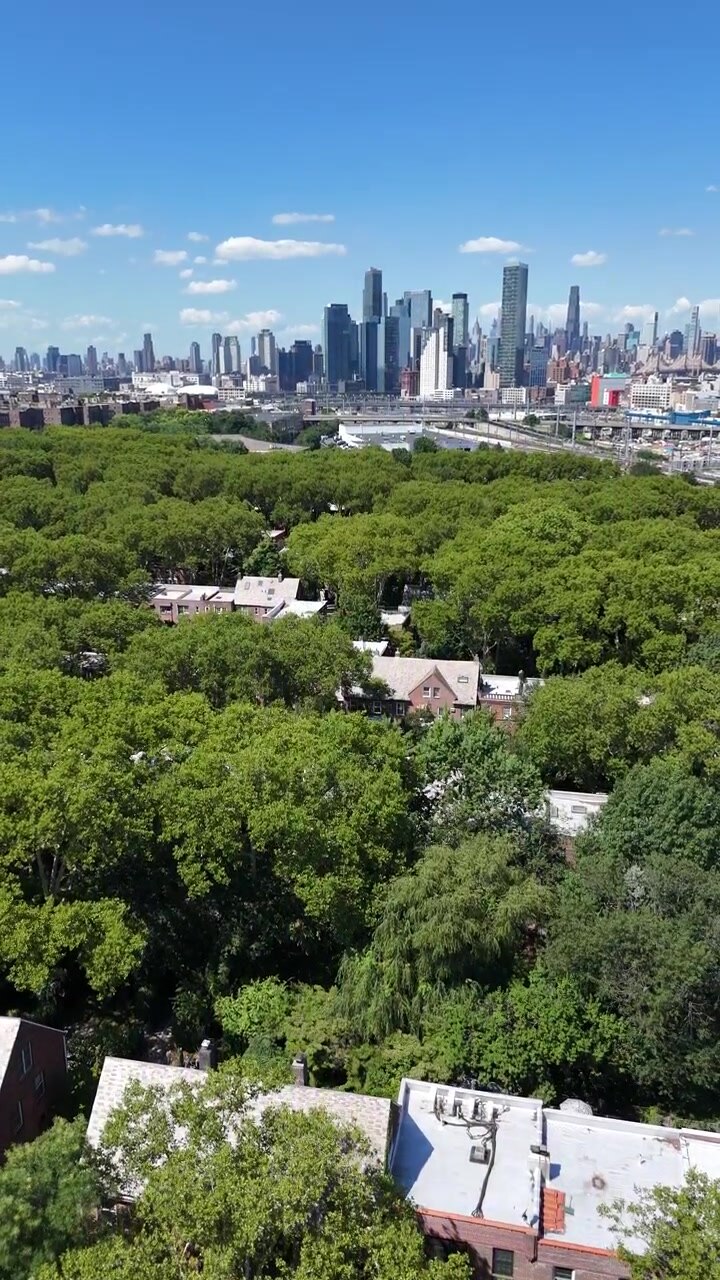 Toma aérea vertical con inclinación ascendente sobre el frondoso dosel arbóreo de Sunnyside Gardens para revelar el horizonte de Midtown Manhattan bajo un cielo azul, Queens, Nueva York, EE. UU.