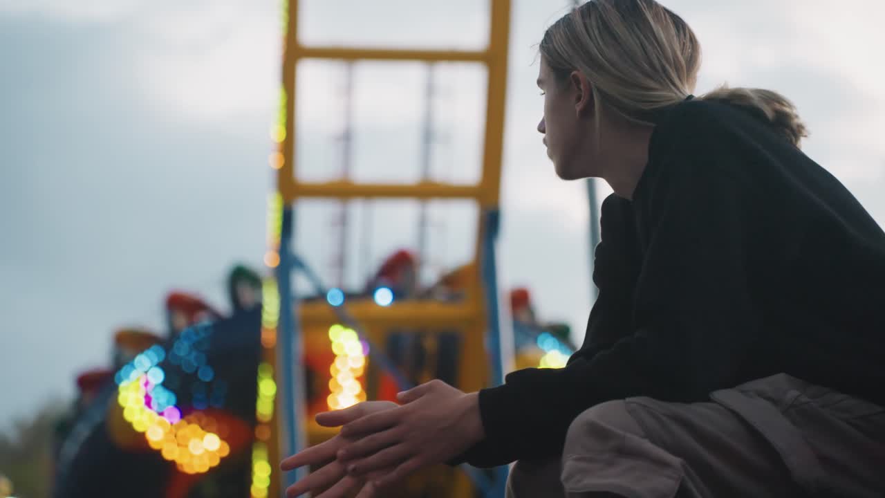 Giovane donna seduta vicino a una giostra, sotto un cielo coperto. Capelli intrecciati raccolti, mani giunte, osserva la ruota illuminata e le luci del luna park. Toni malinconici del crepuscolo, un ritratto di profilo spontaneo che evoca solitudine.