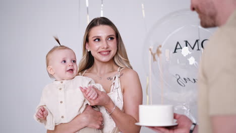 beautiful blonde woman with baby in hands blowing a candle on the cake. smiling man holding a dessert in front of his wife and child.