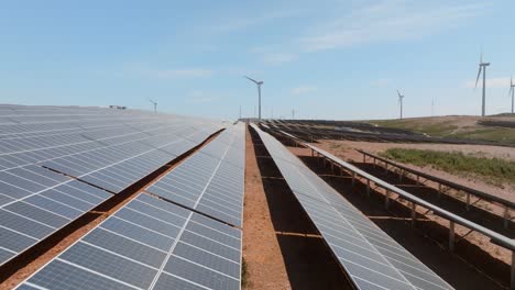 Drohne-Fliegt-Tief-über-Sonnenkollektoren-Im-Bauernhof-In-Richtung-Windkraftanlage-In-Der-Ferne