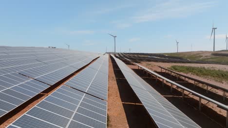 drone volando bajo sobre paneles solares en la granja hacia la turbina eólica en la distancia