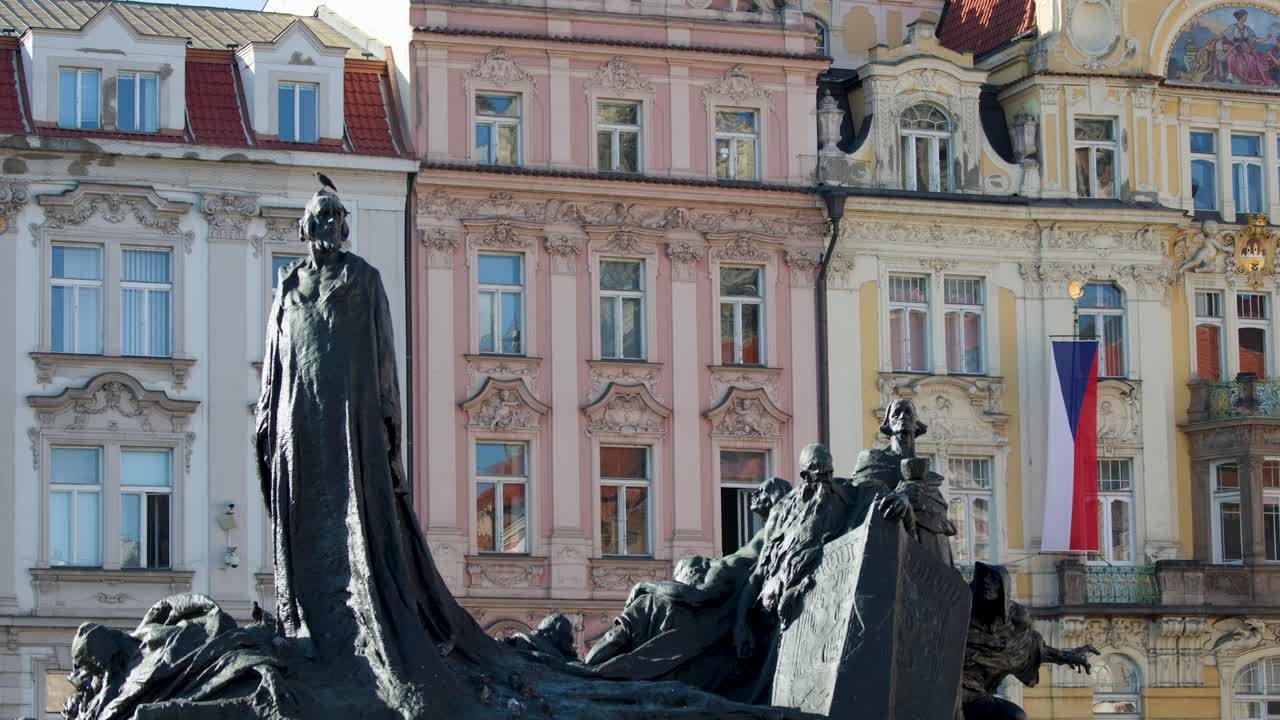 Static shot of Jan Hus Memorial with colorful baroque buildings and Czech flag in daylight