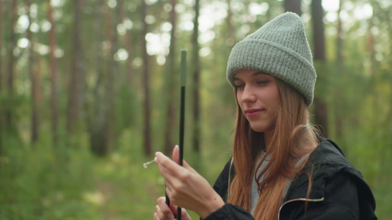 Lady in hoodie and knitted beanie focuses on reinserting rope into tent pole while standing in lush forest, surrounded by tall trees and natural greenery during outdoor camping setup