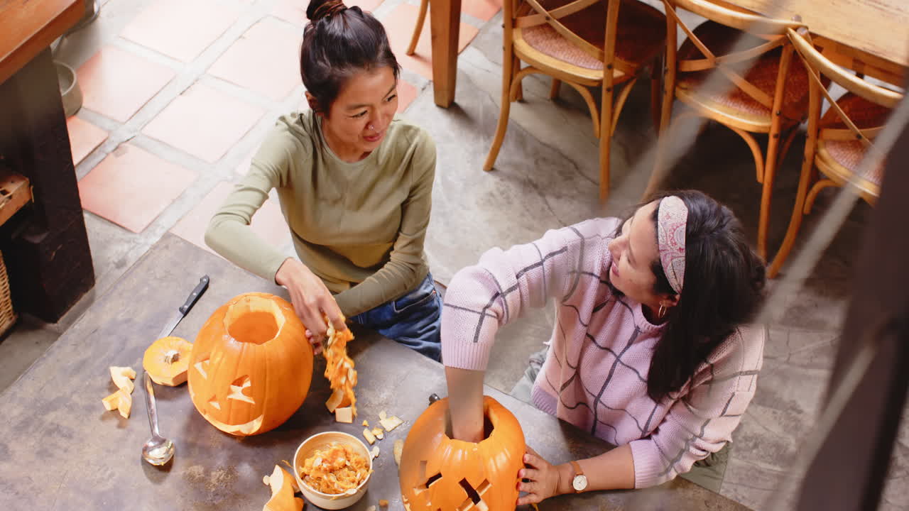 Carving pumpkins together, multiracial grandmother and young woman, at home, Halloween time