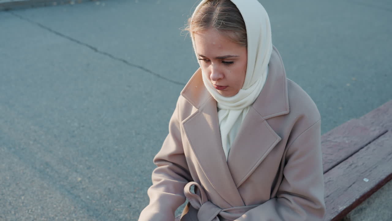 Giovane donna su una panchina che maneggia un medaglione, indossa un foulard e un cappotto di lana, sola in una strada tranquilla, armeggia con la chiusura, espressione introspettiva, luce fredda e soffusa, atmosfera nostalgica cinematografica.