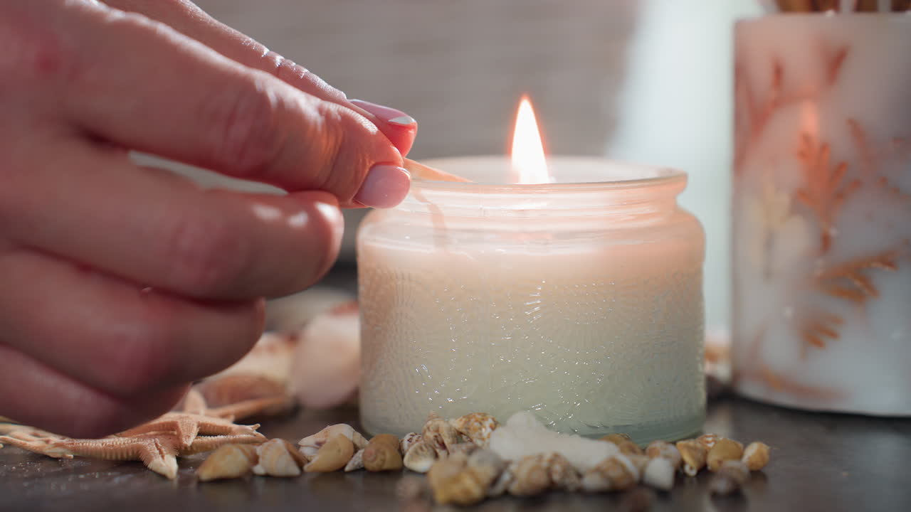 Close up of hand with polished nails lighting white candle with matchstick, flame catching wick surrounded by seashells and starfish on dark marble surface in cozy calm decorative setting