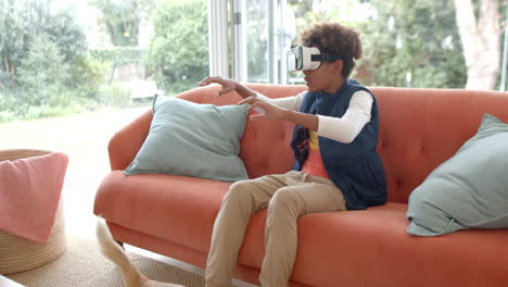 niño afroamericano sentado en el sofá, usando auriculares vr y tocando la pantalla virtual, cámara lenta