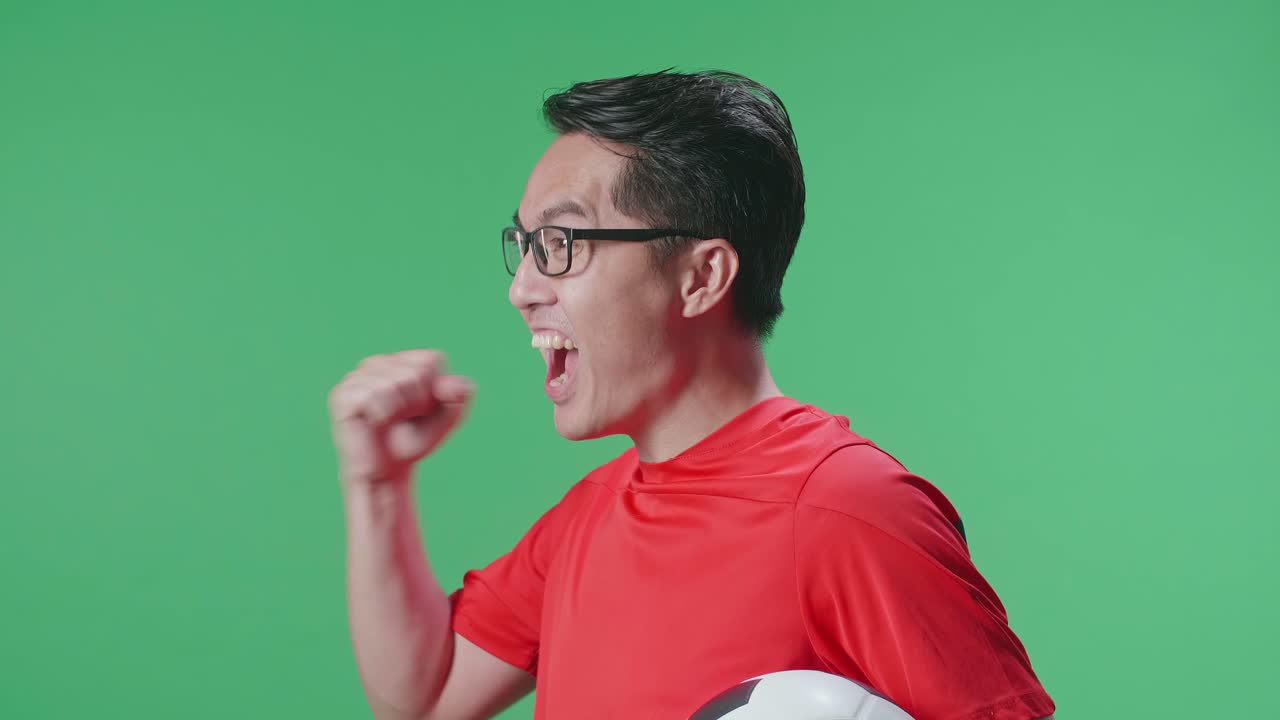 공을 들고 있는 아시아인 남성의 가까운 면 뷰. 골을 외치며 우승팀을 축하하면서 초록색 스크린 배경에서 축구를 응원합니다.