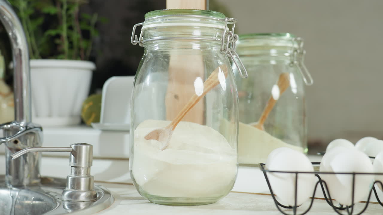 Glass jar filled with milk and wooden spoon placed on clean kitchen counter near sink and egg basket, with mirror reflection showing partial view of someone walking in behind bright kitchen setup