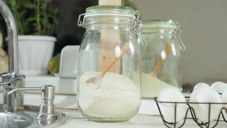 glass jar filled with milk and wooden spoon placed on clean kitchen counter near sink and egg basket, with mirror reflection showing partial view of someone walking in behind bright kitchen setup