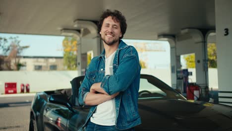 A-young-brunette-guy-in-a-denim-jacket-poses-and-looks-at-the-camera,-he-folded-his-arms-on-his-chest-and-leaned-on-his-dark