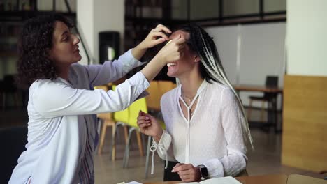 Two-caucasian-women-making-fun-during-their-preparation-for-exams.-Brunette-girl-sticks-yellow-papers-with-painted-eyes-on-her-female-friend's-eyes.-Slow-motion