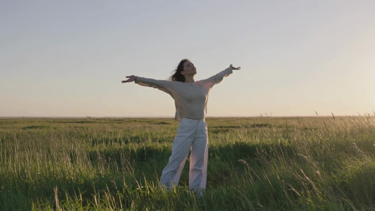 Mujer abriendo los brazos y levantando el rostro mientras el sol desciende en el horizonte derecho, en medio de hierba alta, con pantalones de punto.