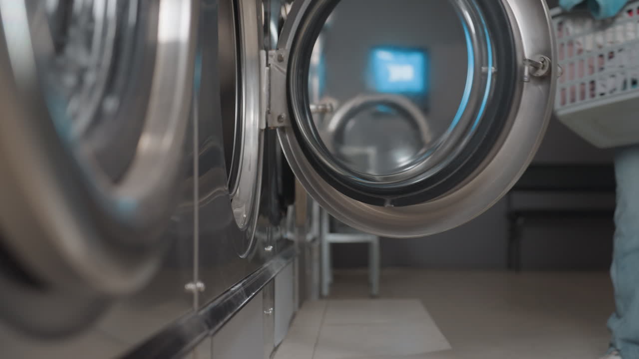 Female hands remove warm towels from dryer into laundry basket inside laundromat, stainless drums row visible, door open as worker unloads fabrics, sorting items before folding, service workflow