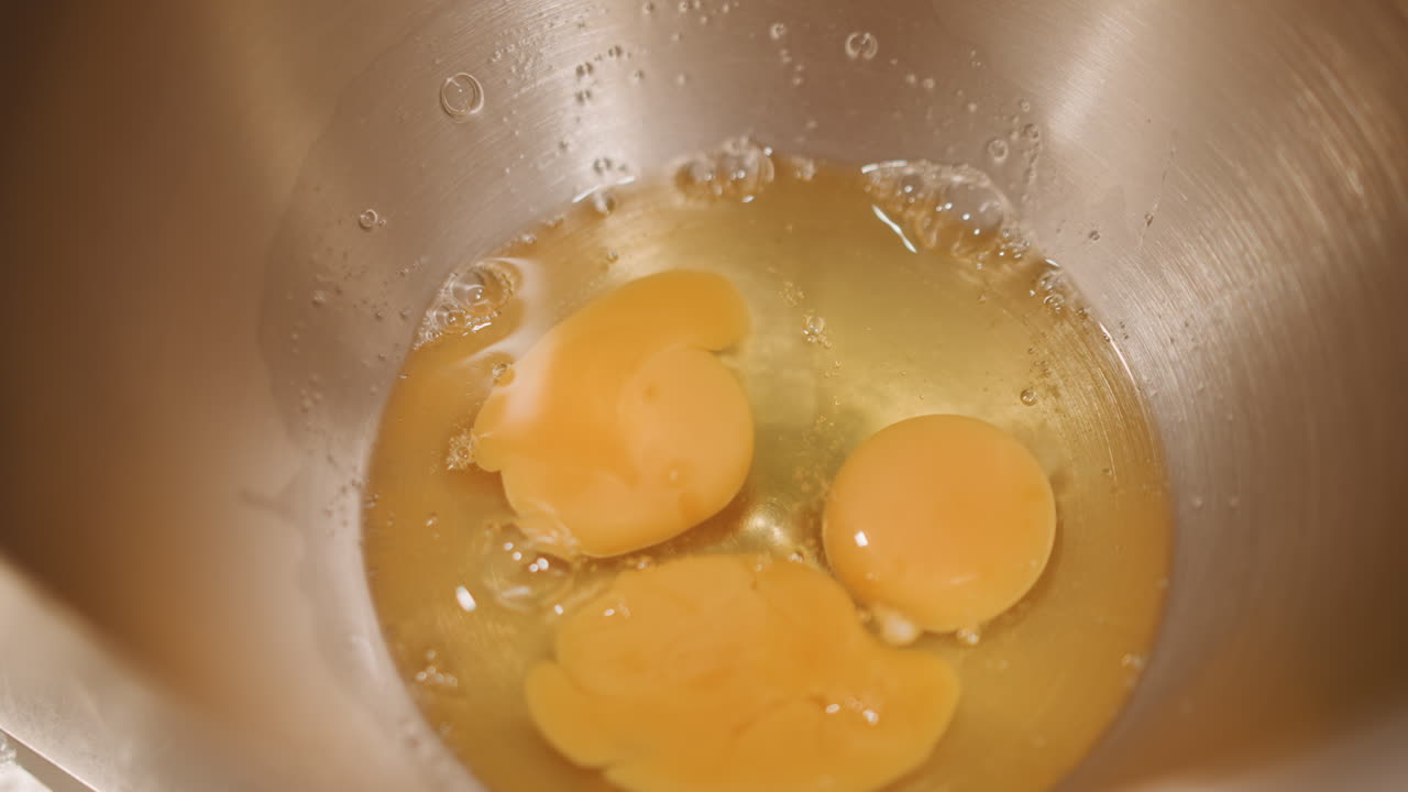 Mangkuk logam dari atas berisi kuning telur, koki pastry memasang mixer untuk isian pai labu, gerakan dekat pengocok, pantulan baja tahan karat, pengukuran yang tepat, dan suasana dapur yang meriah