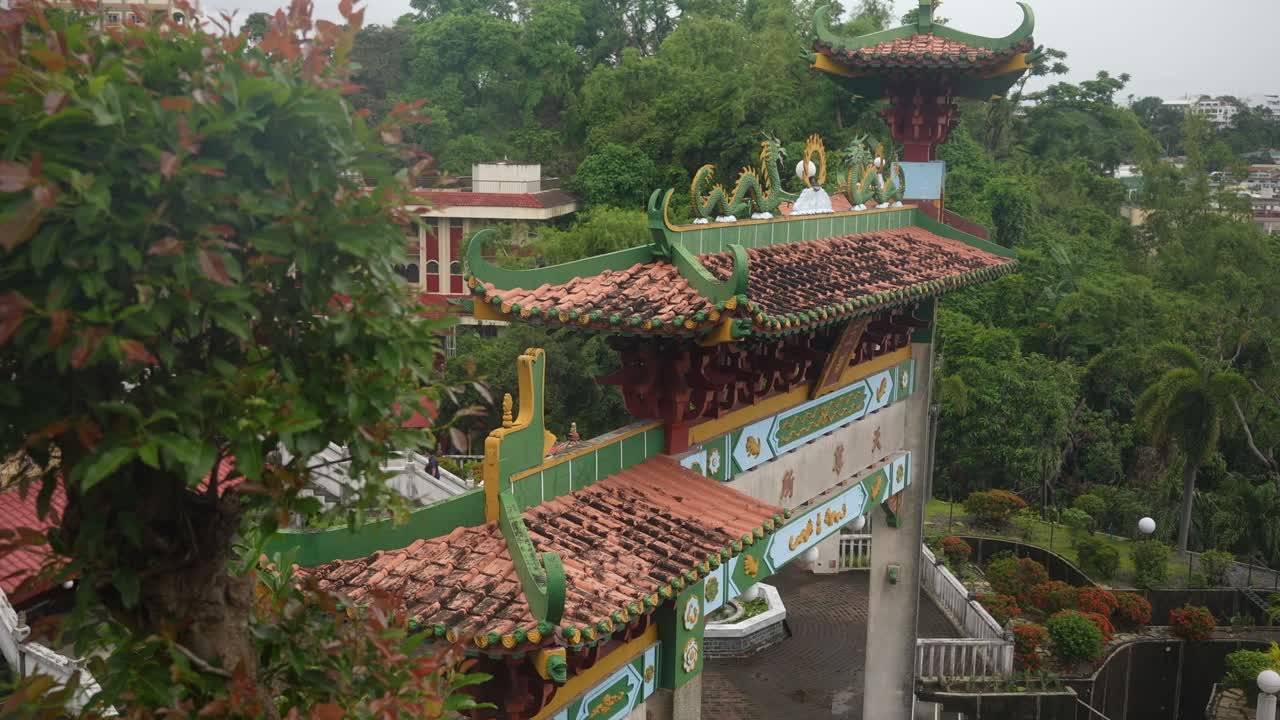 A wide static angle holds throughout, framing a temple gate with ornate curved roofs, vibrant red and green detailing, and symbolic figures. Lush trees and manicured shrubs complete the scene