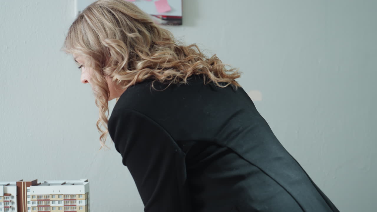Businesswoman walks into brightly lit modern office carrying architectural building model and gently places it on desk, background includes stack of documents