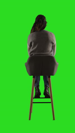 vertical video rear view studio shot of woman sitting on stool facing away from camera against green screen