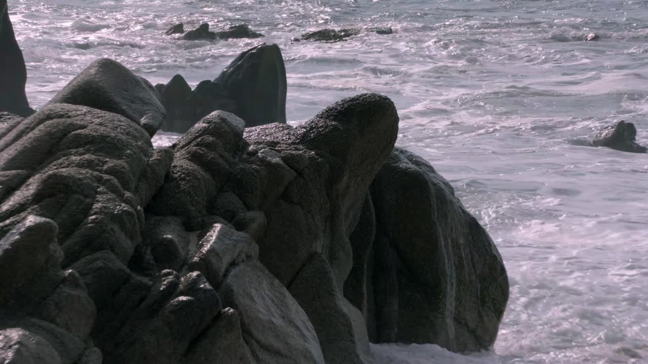 A close-up shot captures ocean waves crashing against rugged coastal rocks, with white foam splashing dramatically in a dynamic seascape scene.
