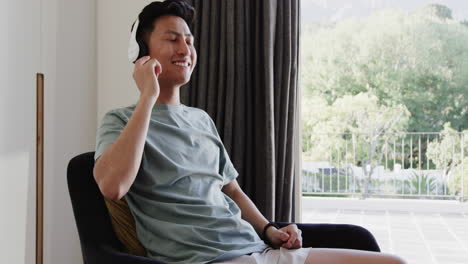 asian man enjoying music with headphones, relaxing at home by window