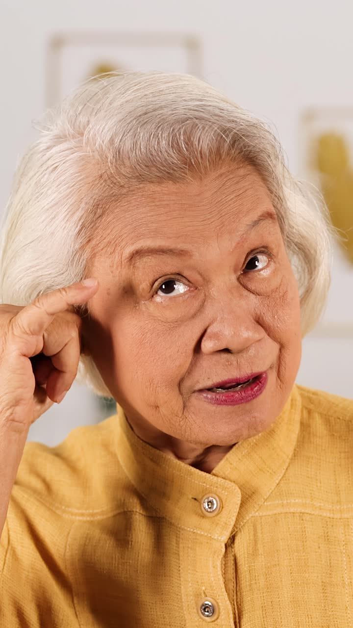 Smiling older Asian woman in yellow shirt points to temple, making playful gesture indoors